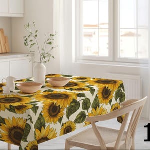 Könnte beinhalten: Ein rechteckiges Tischtuch mit einem Sonnenblumenmuster in Gelb, Grün und Braun. Das Tuch bedeckt einen Holztisch, auf dem ein Holzstuhl und zwei Holzschalen stehen. Eine weiße Vase mit Grün steht auf dem Tisch.