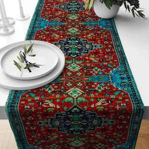 May include: A vibrant red and blue patterned table runner with a detailed, ornate design. The runner features a central red field with intricate teal, navy, and green geometric motifs. A teal border frames the edges. The runner is displayed on a white surface.