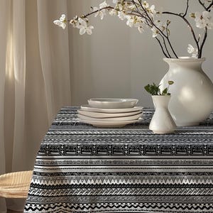May include: A dining table set with a black and white patterned tablecloth, white ceramic plates, and a white vase with flowers. A smaller white vase with greenery is also present. A wooden chair is visible on the left.