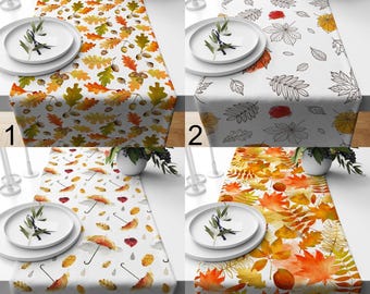May include: Four table runners with fall-themed designs. The first features oak leaves and acorns in orange, yellow, and green. The second has outlined leaves in black and orange on a white background. The third shows umbrellas and falling leaves. The fourth has colorful fall leaves.