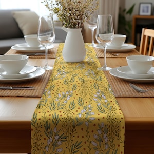 Op de afbeelding: Een gele tafelloper met een bloemenpatroon, met witte bloemen, groene bladeren en bruine accenten. De loper ligt op een houten tafel die gedekt is voor een maaltijd, met witte borden, glazen en een vaas met bloemen.