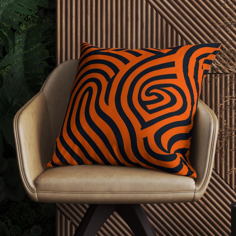 May include: Orange and black patterned throw pillow with a swirling design. The pillow is sitting on a brown leather chair.