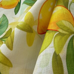May include: Close-up of a fabric with a lemon and floral pattern. The design features bright yellow lemons, green leaves, and white flowers on a cream-colored background. The fabric has a textured appearance.