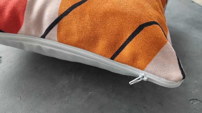 May include: A close-up of a pillow with a zipper closure. The pillow has a geometric pattern in shades of orange, red, and white.