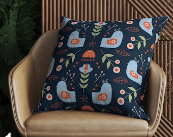 May include: A decorative square pillow with a navy blue background featuring a repeating pattern of light blue birds, orange accents, and green foliage. The pillow is placed on a tan leather chair.