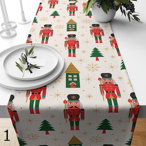 May include: A white table runner with a repeating pattern of nutcrackers, Christmas trees, snowflakes and houses. The nutcrackers are wearing red and green uniforms and holding a staff. The houses are green with gold roofs. The background is a light beige colour.