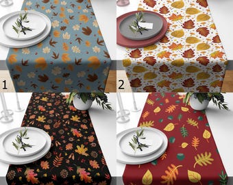 May include: Four table runners with autumn leaf patterns are shown. The runners have different colour schemes: blue, white, black, and red. Each runner is decorated with plates, greenery, and candles, creating a seasonal table setting.
