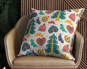 May include: A decorative pillow with a festive Christmas pattern. The pillow features a white background with colorful illustrations of Christmas trees, ornaments, hearts, and other holiday-themed designs. The pillow is placed on a tan chair.