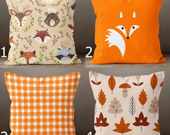 May include: Four decorative throw pillows with autumn-themed designs. One pillow features woodland animal faces, another has a white fox on an orange background, a third has an orange and white checkered pattern, and the last has fall leaves.