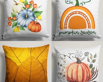 May include: Four white throw pillows with autumn-themed designs. One pillow features a watercolor arrangement of pumpkins and foliage. Another displays an orange rainbow with the text "pumpkin season." The third has a close-up of yellow leaves. The last pillow has a watercolor pumpkin.