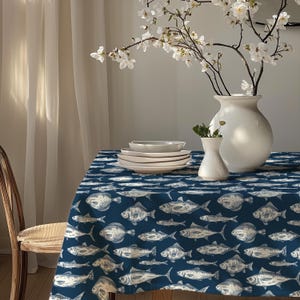 May include: A dark blue tablecloth featuring a repeating pattern of white fish illustrations. On the table, there is a stack of white ceramic plates, a small white ceramic vase, and a large white ceramic vase containing white flowers.