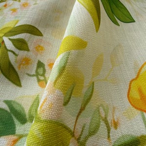 May include: Close-up of a folded fabric with a floral pattern. The design features large yellow flowers with orange accents, green leaves, and small white and pink blossoms on a cream-colored background. The fabric has a textured appearance.
