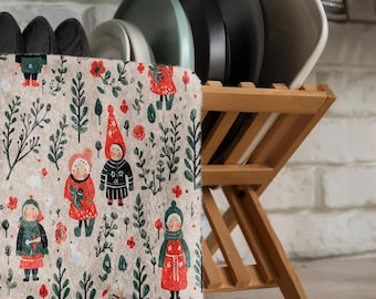 May include: A beige kitchen towel with a festive pattern of red and green figures in a winter forest setting. The towel is hanging on a wooden dish rack with a stack of plates.