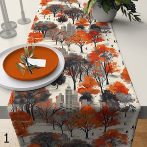 Puede incluir: Un camino de mesa con un diseño de temática otoñal. El camino de mesa presenta un paisaje urbano con árboles en tonos naranja, gris y negro. El diseño incluye siluetas de personas caminando en un entorno similar a un parque. Un plato naranja y un plato blanco están sobre la mesa.