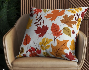 Op de afbeelding: Een vierkant decoratief kussen met een patroon van herfstbladeren in rood, oranje en geel op een witte achtergrond. Het kussen ligt op een bruine leren stoel, met een houten lattenwand op de achtergrond.