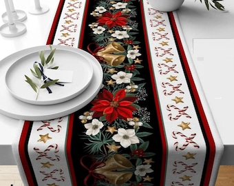 May include: A Christmas-themed table runner with a black center adorned with red poinsettias, white flowers, gold bells, and stars. The runner has red and white borders with candy cane and star patterns. A white plate with greenery sits on the table.