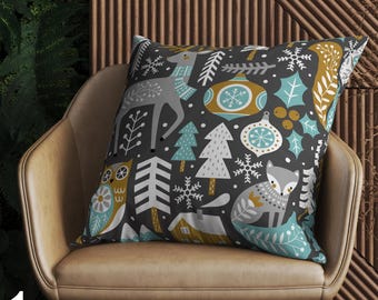 May include: A cushion with a dark grey background, decorated with a winter-themed pattern. The design includes a deer, fox, owl, snowflakes, Christmas ornaments, and trees in grey, teal, and gold. The cushion is placed on a tan chair.