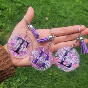 May include: Three purple and white glitter keychain charms with the names Tori, Brianna, and Leah written in white with a black initial letter.