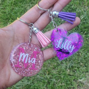 May include: Two glitter resin keychains with tassel charms. One keychain is pink with the name "Mia" and the other is purple with the name "Lilly".