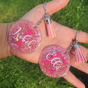 May include: Two pink glitter resin keyrings with silver key rings and pink tassels. One keyring says "Isabel" and the other says "Ella".