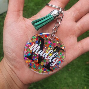 May include: A round, clear keyring with a colourful glitter design and the name "Maddy" written in white. The keyring has a green tassel attached to a silver ring.
