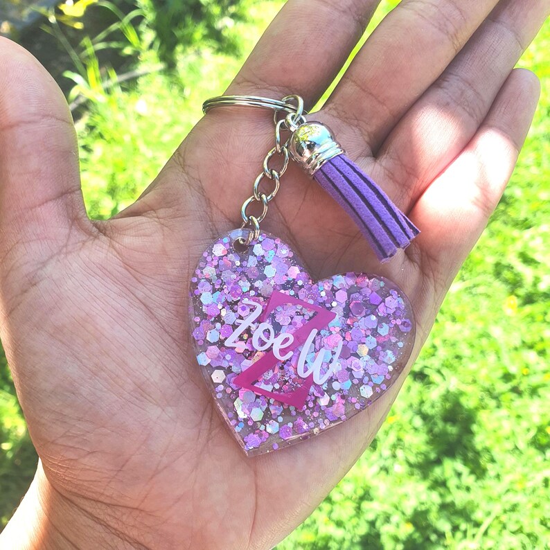 May include: A pink heart-shaped keyring with glitter embedded in the resin. The keyring has a silver key ring and a purple tassel. The name "Zoe" is written in pink letters on the heart.