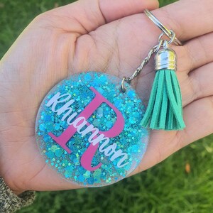 May include: A teal blue glitter keychain with a pink letter R and the name "Rhiannon" written in white. The keychain has a silver chain and a teal blue tassel.