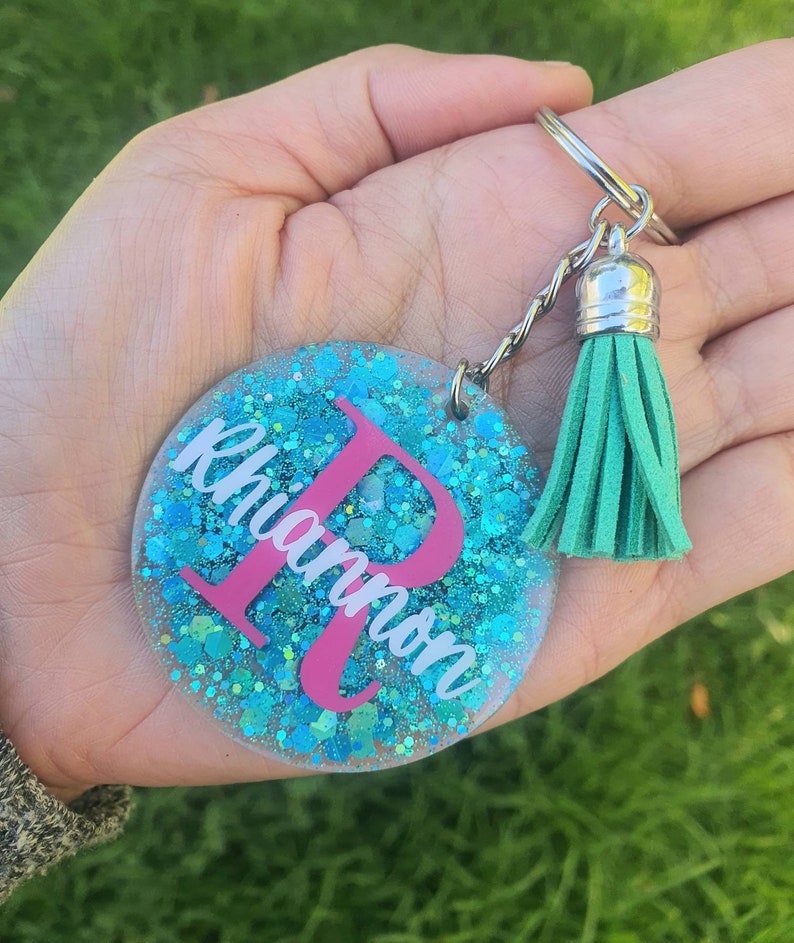 May include: A teal blue glitter keychain with a pink letter R and the name "Rhiannon" written in white. The keychain has a silver chain and a teal blue tassel.