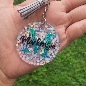 May include: A round, clear keyring with a silver chain and tassel. The keyring has a sparkly, iridescent background with a teal letter "M" and the name "Mackenzie" written in black.