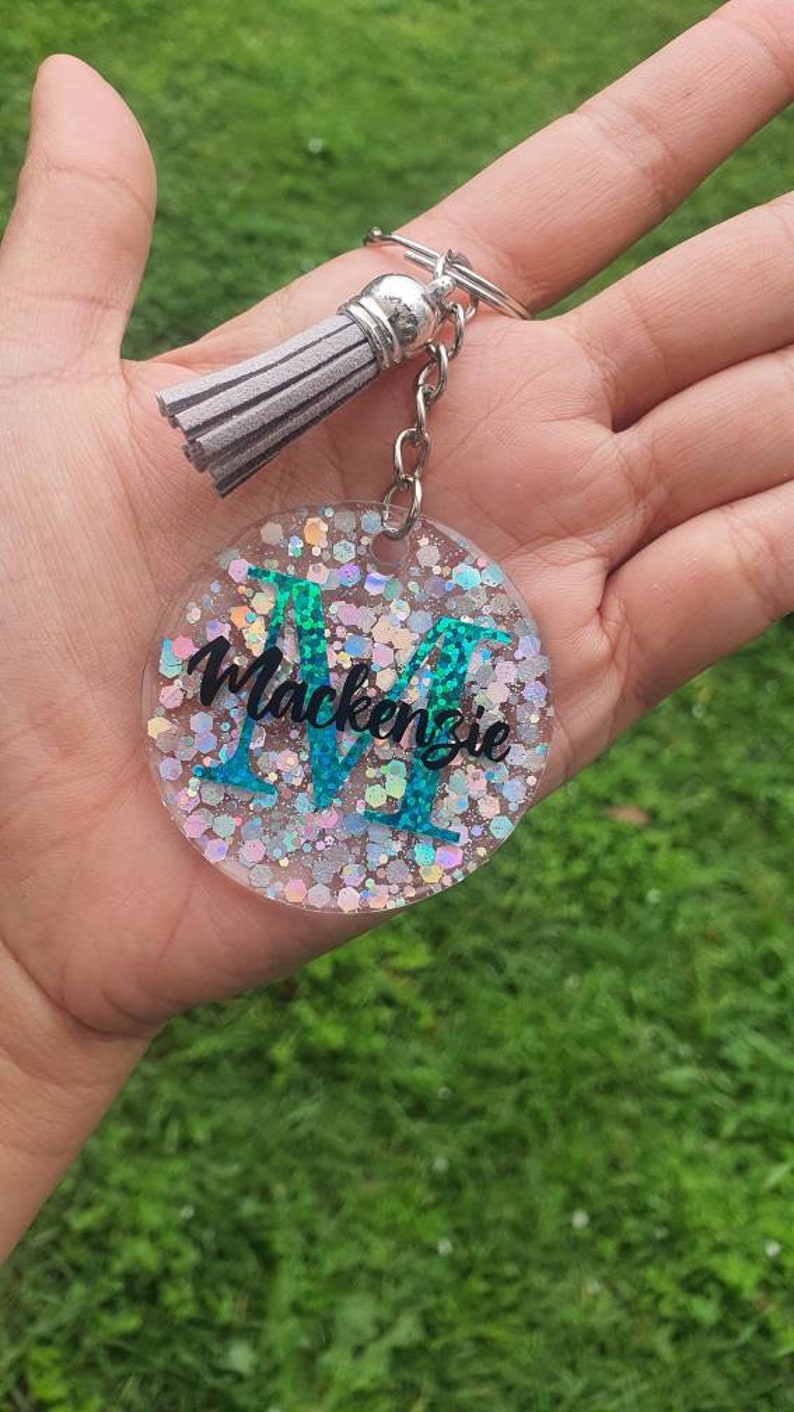 May include: A round, clear keyring with a silver chain and tassel. The keyring has a sparkly, iridescent background with a teal letter "M" and the name "Mackenzie" written in black.