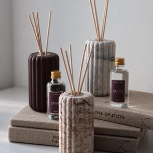 May include: Three cylindrical reed diffusers with fluted textures in dark brown, white marble, and beige swirl patterns. Each diffuser holds light-coloured wooden reeds. Two clear glass bottles with gold caps and labels reading ORIGIN are included.