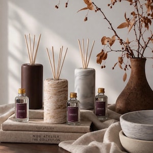 May include: Three cylindrical reed diffusers in solid brown, beige marble, and grey marble finishes, each with wooden reeds. Three small clear glass bottles with gold caps and labels reading ORIGIN, placed on two stacked books labelled NOU ATELIER.