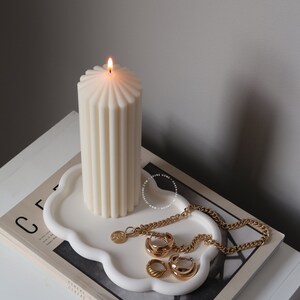 May include: A white ceramic tray with a cloud-like shape, featuring a lit white pillar candle, a gold chain necklace, and gold hoop earrings. The tray is placed on a magazine with the text "Cereal" printed on the cover.