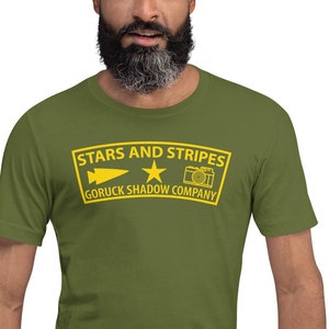 May include: A green t-shirt with a yellow patch that says "Stars and Stripes Goruck Shadow Company" with a star, a camera, and a lightning bolt.