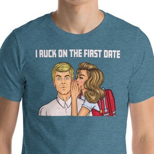 May include: A teal blue t-shirt with the text "I RUCK ON THE FIRST DATE" in white. The shirt features a pop art style illustration of a woman whispering in a man's ear.