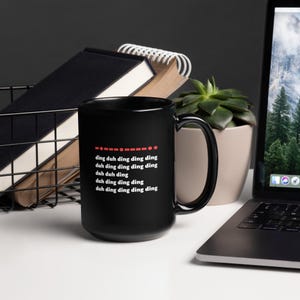 May include: A black ceramic mug with the text "ding duh ding ding ding duh ding ding ding ding duh duh ding duh ding ding ding duh ding ding ding ding" in white. The mug is on a white surface with a laptop and a succulent plant in the background.