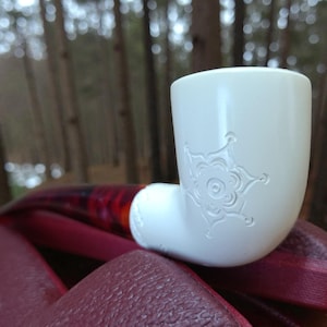 May include: A white ceramic pipe with a floral design etched on the bowl. The pipe is resting on a burgundy surface, with a blurred forest background. The pipe is a smoking accessory.
