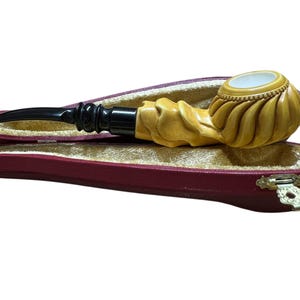 Pode incluir: Um cachimbo de tabaco esculpido com um design em espiral, repousando em um estojo vermelho aberto com um fecho dourado. O cachimbo apresenta um bocal preto e uma tigela de cor clara.