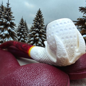 Könnte beinhalten: Nahaufnahme einer Tabakpfeife mit einem weißen Kopf mit einem strukturierten Muster aus kleinen Kreisen. Der Stiel ist rot mit einem gelben Band. Die Pfeife liegt auf einer weinroten Oberfläche, mit einem zweiten weinroten Objekt im Vordergrund. Verschneite Bäume im Hintergrund.