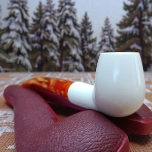 May include: A white tobacco pipe with a red and orange stem rests on a burgundy leather case. The pipe has a rounded bowl and a curved stem. The background features a blurred image of snow-covered pine trees.
