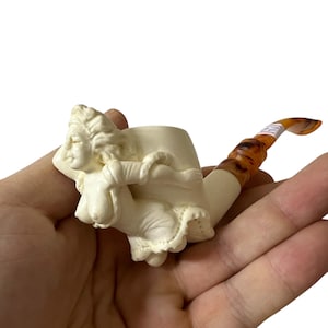 Könnte beinhalten: Elfenbeinfarbene Tabakpfeife mit einer skulptierten, liegenden Frauenfigur. Die Pfeife hat einen gebogenen Stiel mit einem marmorierten bernsteinfarbenen und braunen Muster. Ein einzigartiges Sammlerstück.