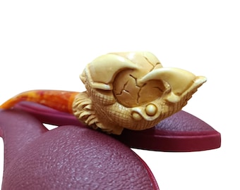 May include: A carved tobacco pipe with a detailed, cream-colored bowl shaped like a mythical creature. The stem is orange and brown, and the pipe rests on a burgundy surface. The pipe is a unique collectible.