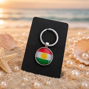 May include: A circular metal keychain featuring the flag of Kurdistan, which consists of horizontal red, white, and green stripes with a central yellow sun emblem, attached to a silver split ring and mounted on a black rectangular display card.