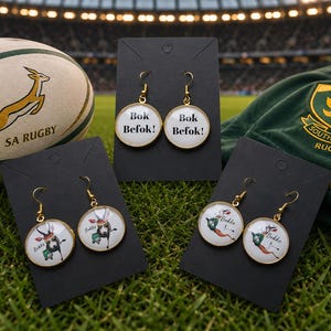 May include: Three pairs of round dangle earrings on black display cards. One pair features the text "Bok Befok!" in black font. The other two pairs feature a cartoon springbok illustration with the text "Bokke" in a gold-toned metal setting.