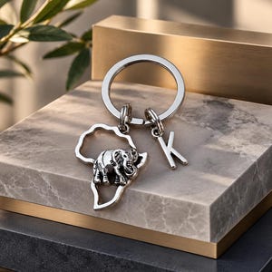 Könnte beinhalten: Ein silberfarbener Metallschlüsselanhänger mit einem runden Ring, einem ausgeschnittenen Anhänger in Form des afrikanischen Kontinents mit einer detaillierten Elefantenfigur darin und einem separaten Anhänger in Form des Großbuchstabens K.
