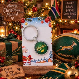 May include: A circular metal keychain featuring a green background with the gold text Bokke and a small gold rugby ball graphic, attached to a silver ring and presented on a card illustrated with a Santa Claus figure and a winter landscape.