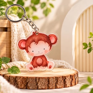 May include: A clear acrylic keyring featuring a printed illustration of a brown monkey with large ears, a round face, and a light-coloured belly. The monkey is in a seated position and is attached to a silver-toned metal split ring by a short chain.