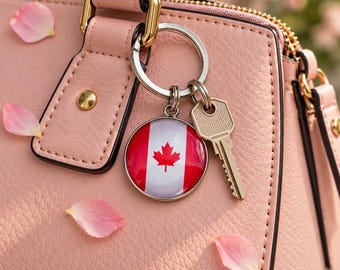 May include: A silver metal keychain featuring a circular charm with the red and white Canadian flag design, including a red maple leaf in the center. The keychain is attached to a silver metal key and hangs from the handle of a light pink handbag.