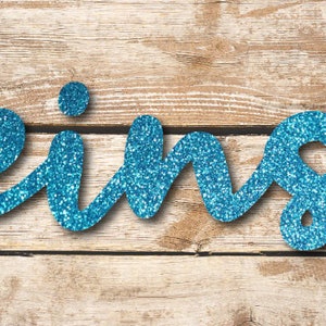 Könnte beinhalten: Das Wort "eins" in funkelnden, blauen Glitzerbuchstaben auf einem Holzgrund.