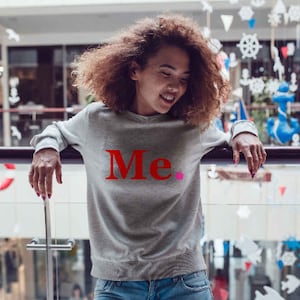 Könnte beinhalten: Eine Frau trägt ein graues Sweatshirt mit dem Wort "Me." in Rot auf der Vorderseite. Sie steht vor einem Geländer und lächelt.